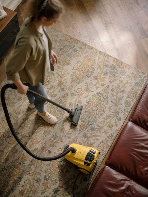 A yellow canister vacuum cleaner with a black hose and wand is positioned on an ornate, multicolored area rug on a wooden floor in a living room. To the left, there is a wooden sideboard with a black